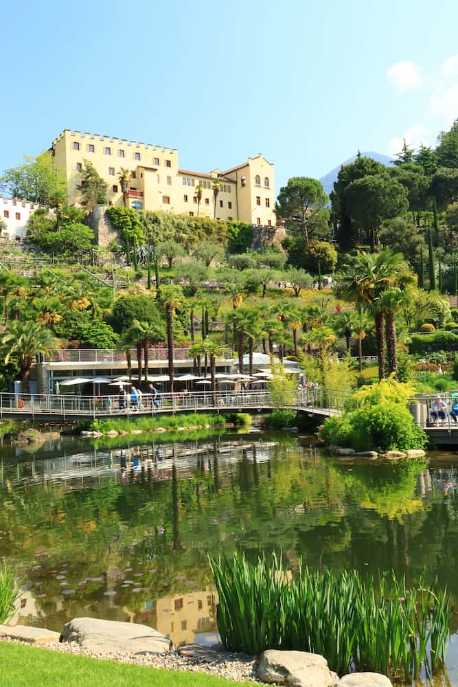 Veduta panoramica dei Giardini Trauttmansdorff di Merano in primavera con lo stagno delle ninfee, il castello e le montagne innevate sullo sfondo.