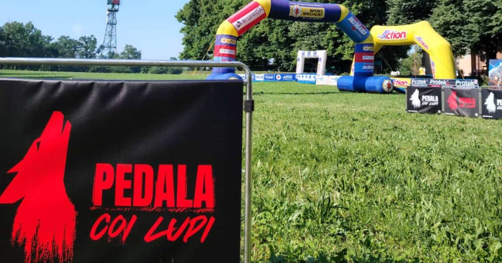 Logo della Pedala coi Lupi in primo piano con l'arco di partenza a Cascina San Fedele nel Parco di Monza.
